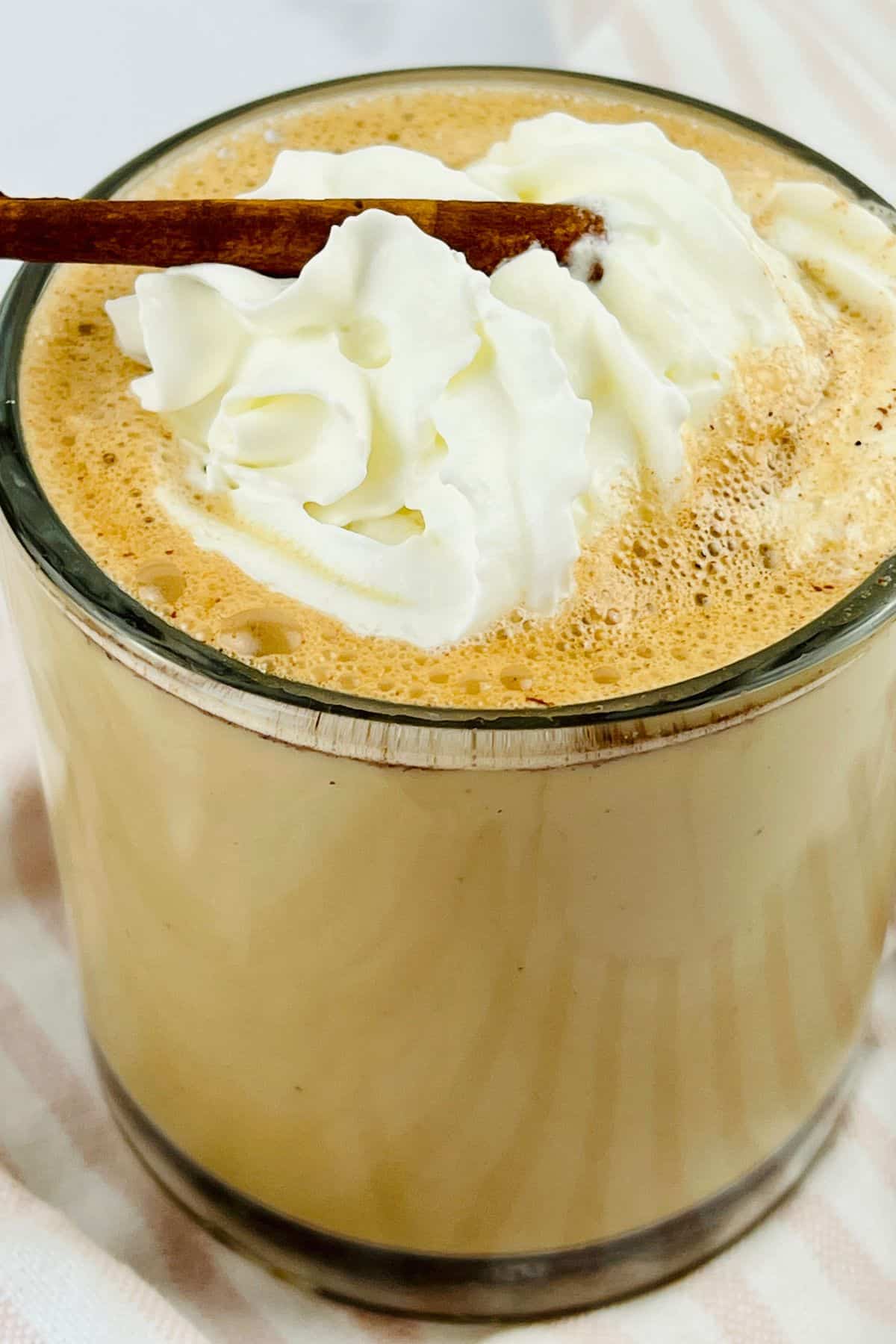 A glass of frothy coffee topped with whipped cream and garnished with a cinnamon stick.