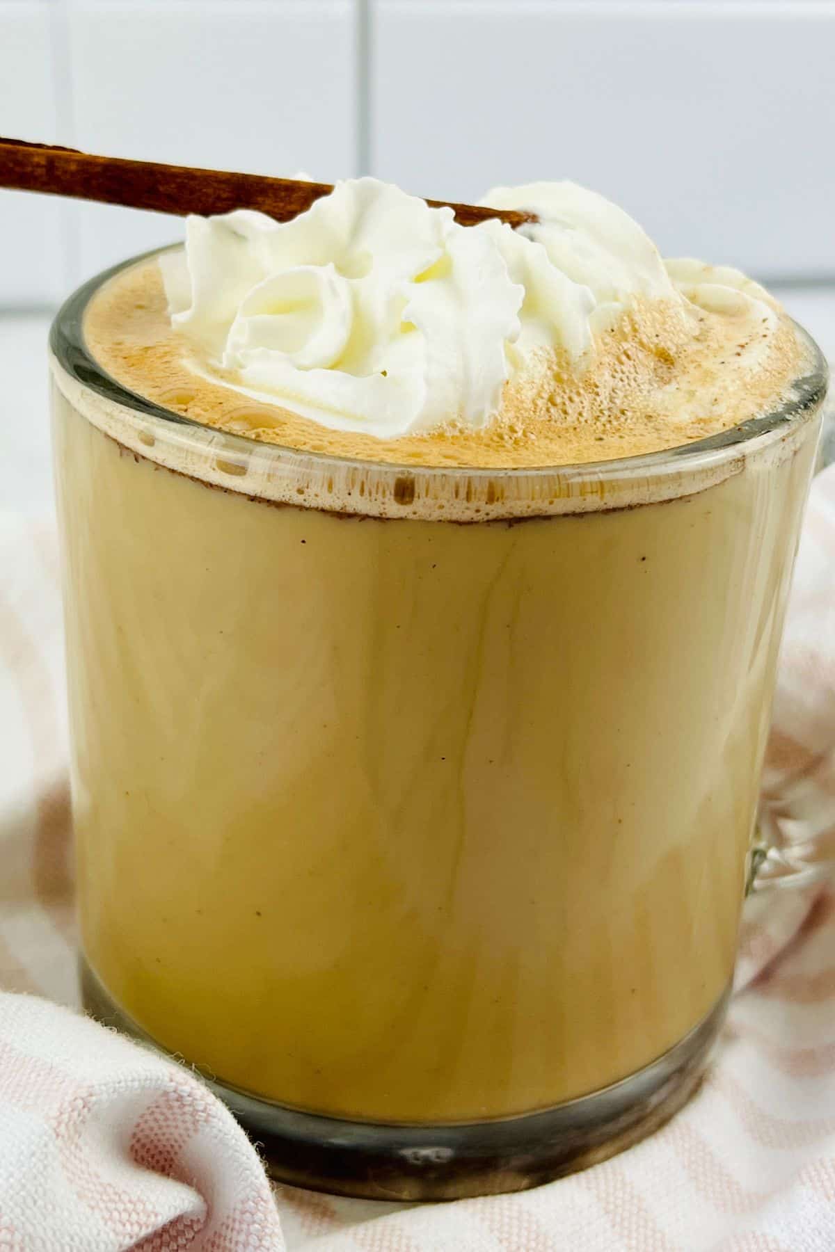 A glass mug filled topped with whipped cream and garnished with a cinnamon stick, sits on a striped cloth.