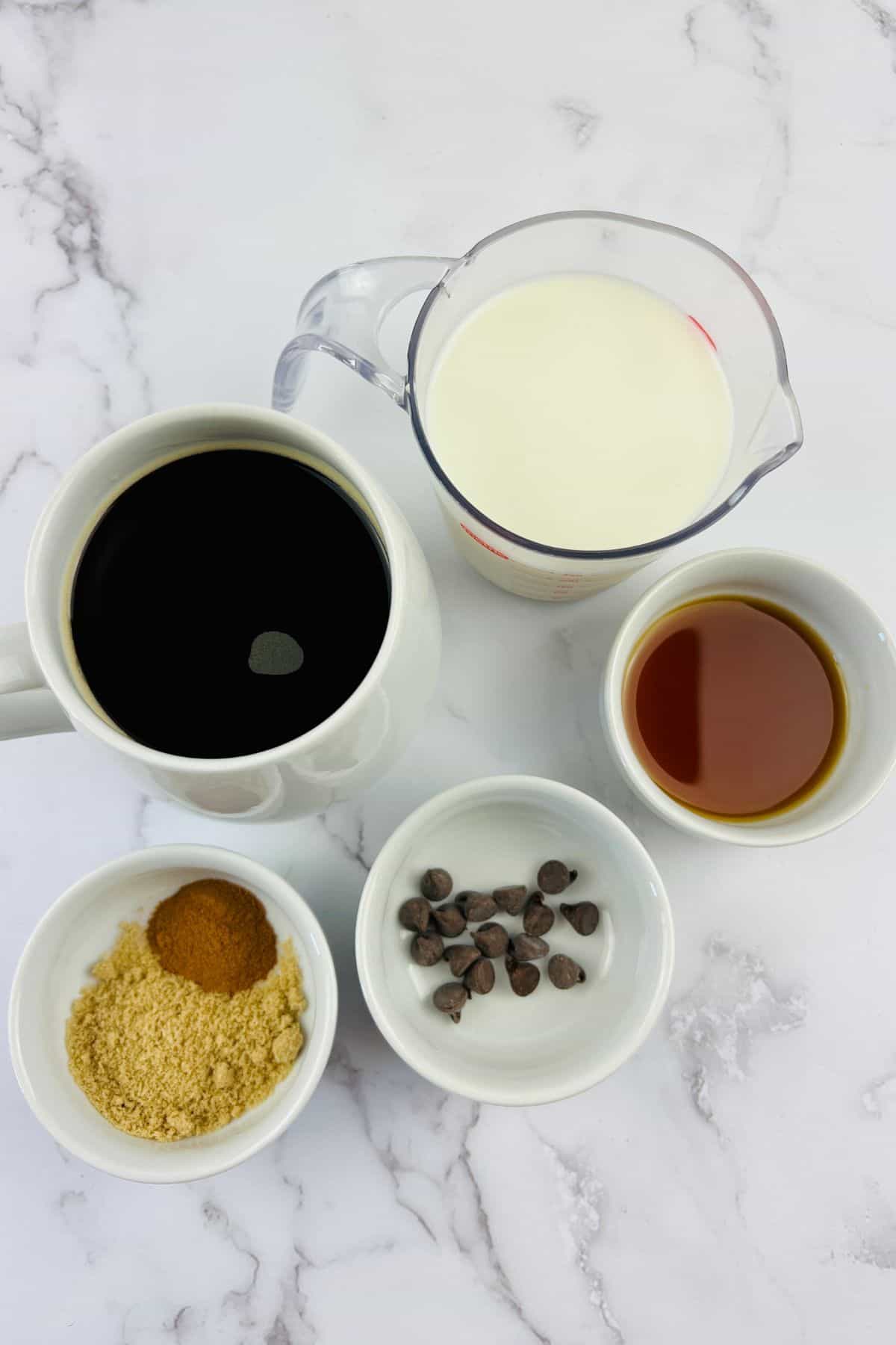 A mug of black coffee, a measuring cup of cream, a small bowl of brown sugar and cinnamon, chocolate chips, and vanilla extract on a marble surface.