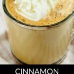 A glass mug filled with a Cinnamon Chocolate Latte, topped with whipped cream and a cinnamon stick, rests on a light-colored surface.