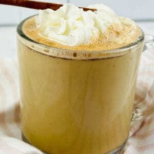 A clear glass mug filled with a frothy Cinnamon Chocolate Latte, topped with whipped cream and a cinnamon stick, rests on a striped cloth.