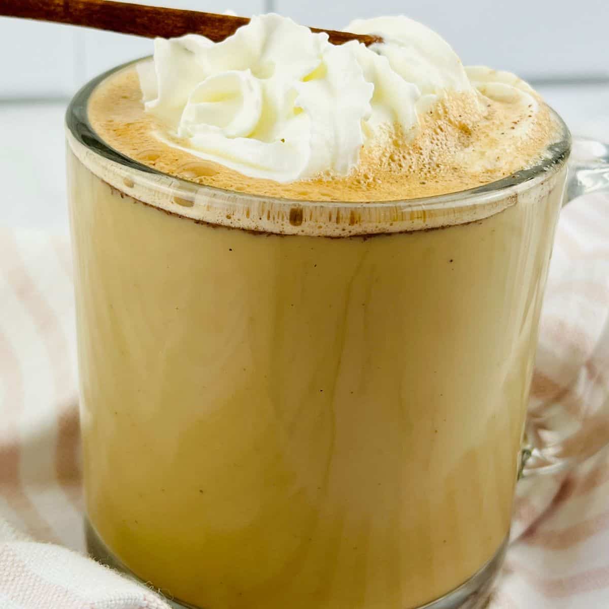 A clear glass mug filled with a frothy Cinnamon Chocolate Latte, topped with whipped cream and a cinnamon stick, rests on a striped cloth.