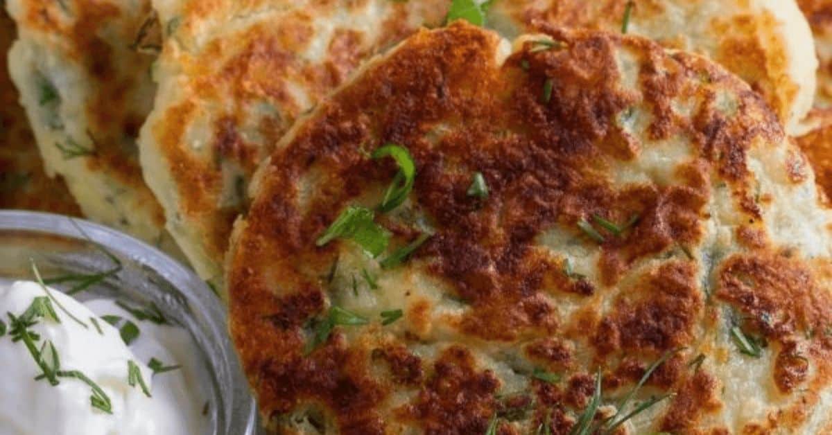 Three golden-brown Fried Mashed Potato Cakes garnished with herbs are served next to a small dish of sour cream.