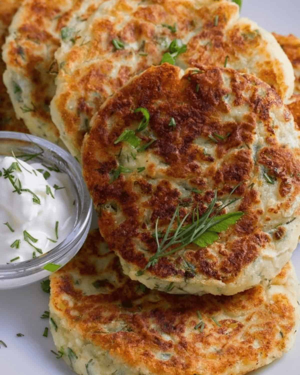 Golden brown Fried Mashed Potato Cakes garnished with fresh dill, served with a side of sour cream in a small glass bowl.