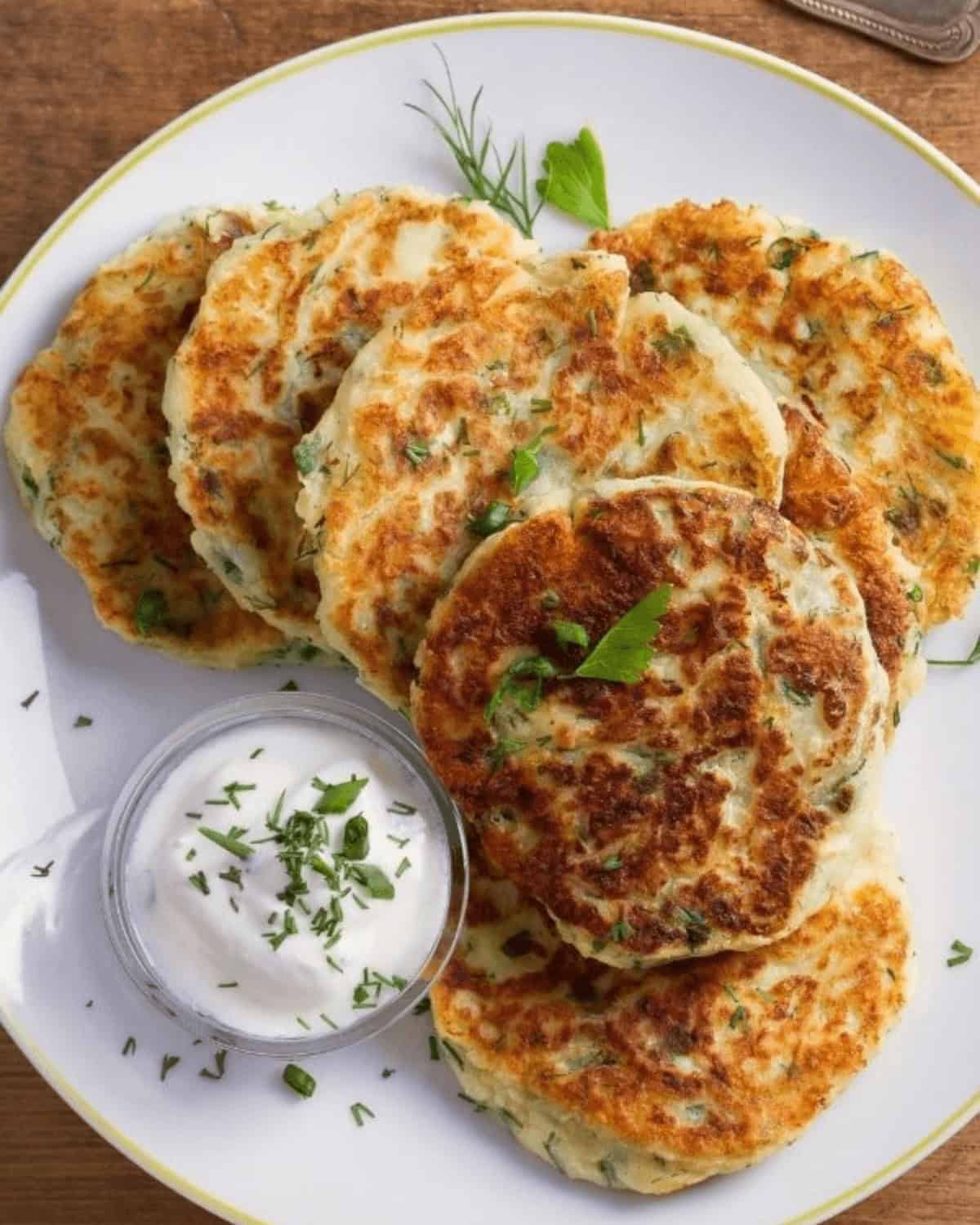A plate of golden-brown fried mashed potato cakes garnished with herbs is served with a small glass bowl of sour cream.