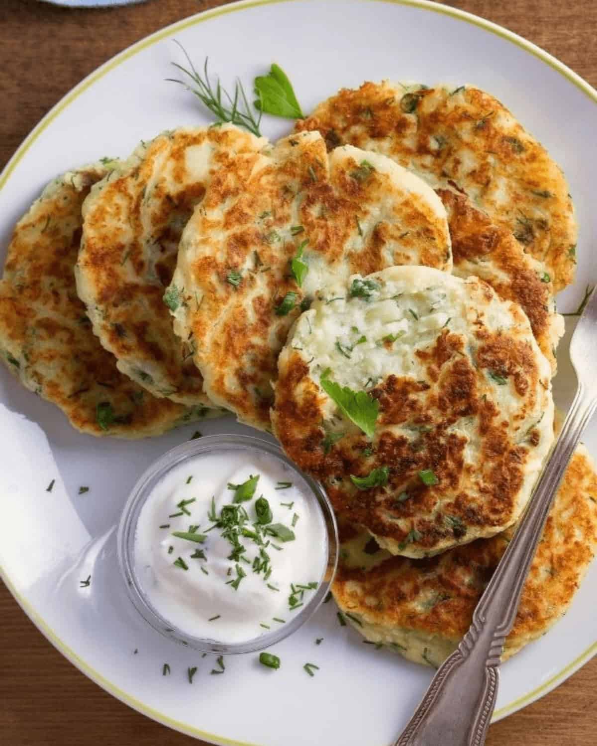 A plate of golden brown Fried Mashed Potato Cakes garnished with herbs, served with a small bowl of creamy dip and a spoon on the side.