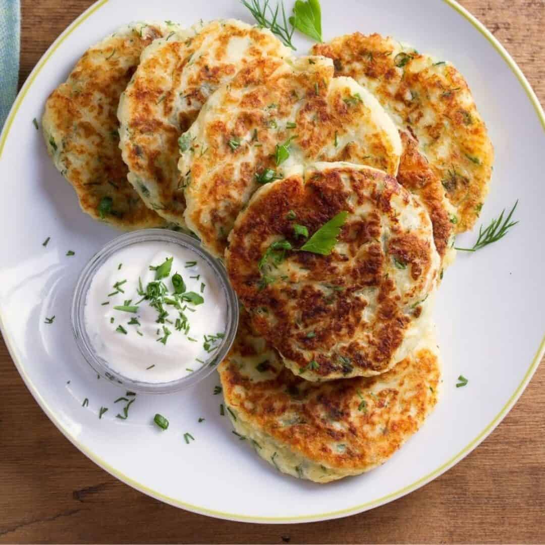 A plate of golden-brown Fried Mashed Potato Cakes garnished with herbs, served with a small dish of sour cream topped with chopped herbs.
