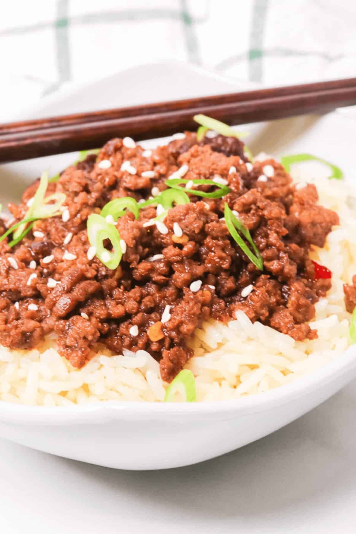 A Beef Bulgogi Bowl features a bowl of white rice topped with seasoned ground meat, garnished with sliced green onions and sesame seeds, with chopsticks in the background.