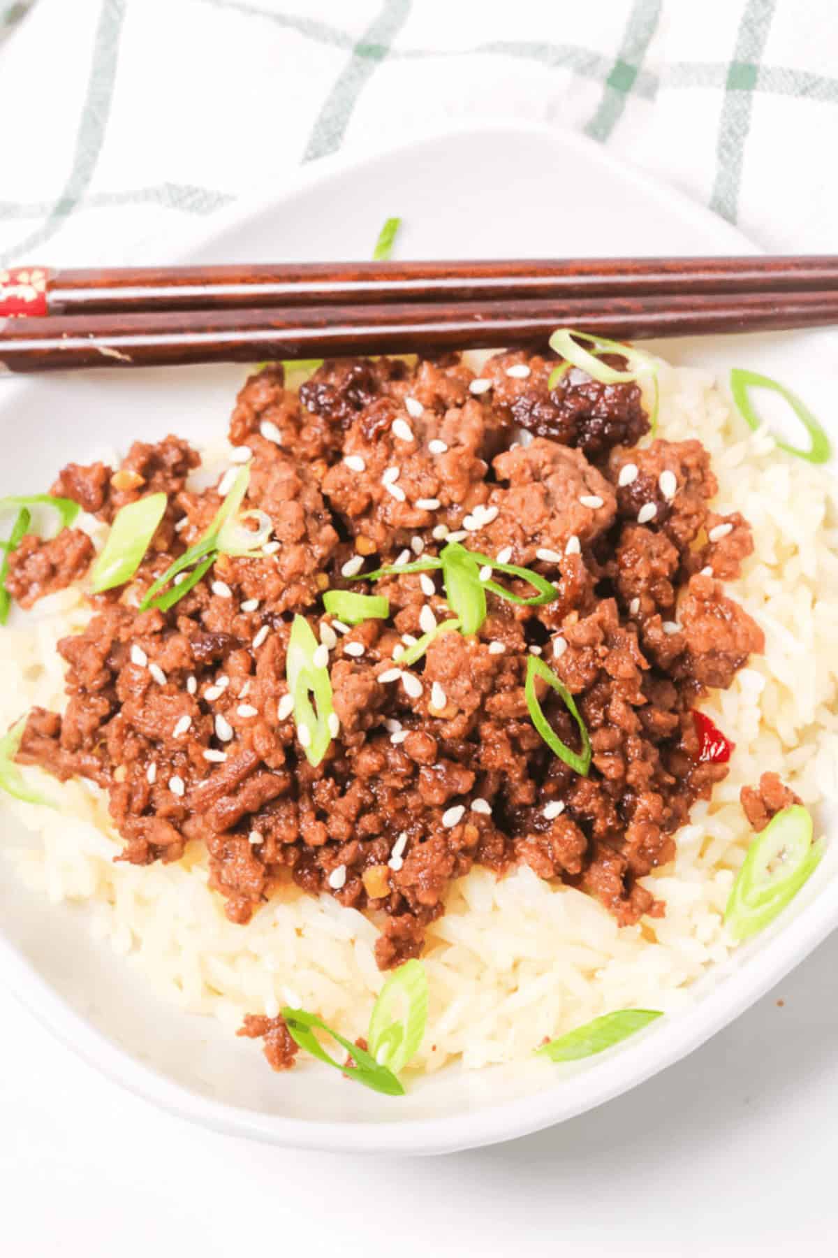 A Beef Bulgogi Bowl featuring a bed of rice crowned with seasoned ground beef, garnished with sliced green onions and sesame seeds, served alongside chopsticks.