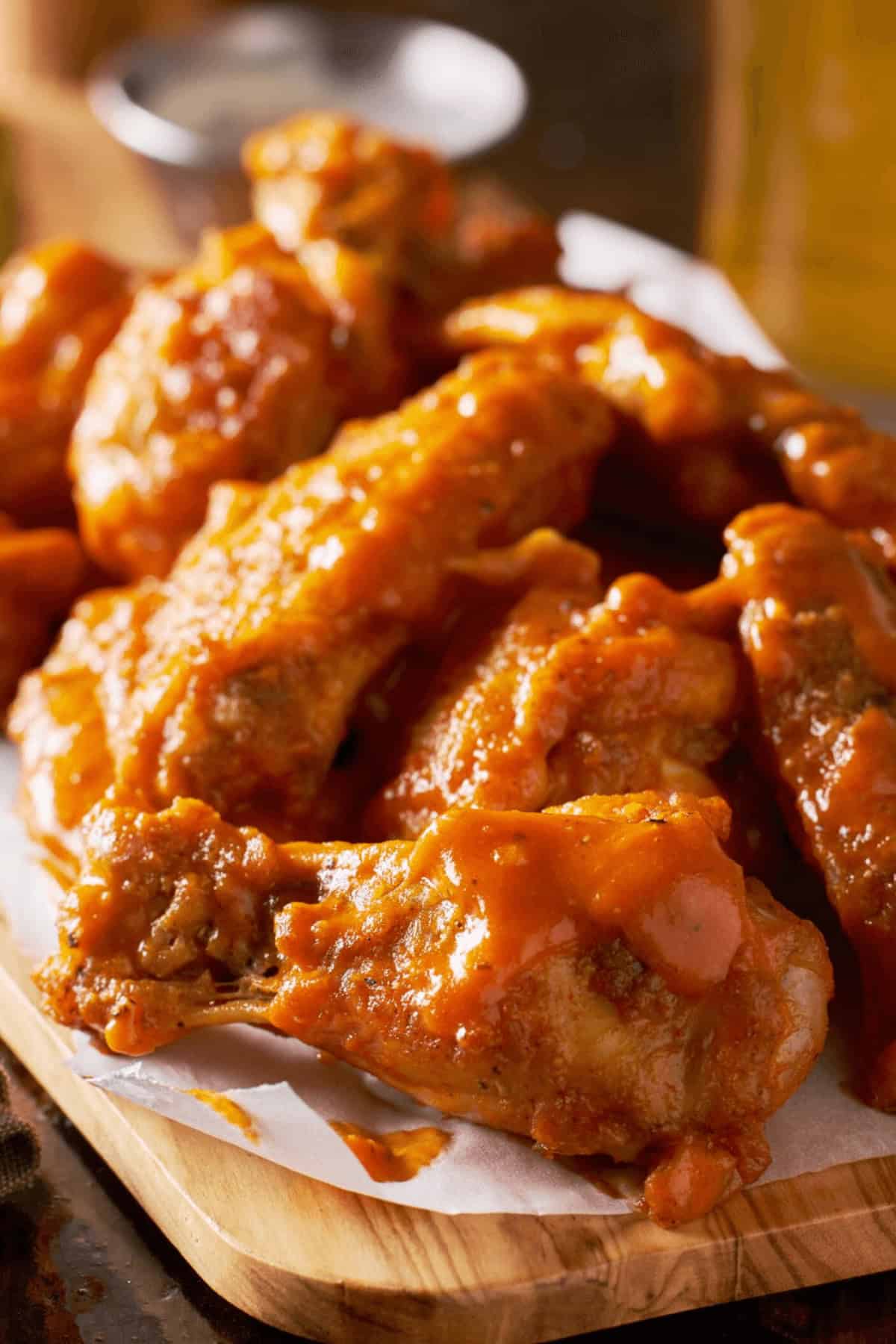 A wooden platter piled with glazed chicken drumsticks, drizzled with a tangy chicken wing sauce, rests on parchment paper.