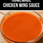 A glass bowl, brimming with bright orange chicken wing sauce, rests on a wooden surface.