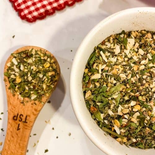A wooden teaspoon and a small white bowl filled with an Italian Seasoning substitute herb and spice mix are on a white surface next to a red and white checkered cloth.