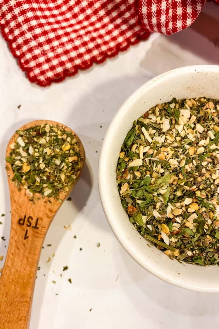 A wooden teaspoon and a small white bowl filled with an Italian Seasoning substitute herb and spice mix are on a white surface next to a red and white checkered cloth.