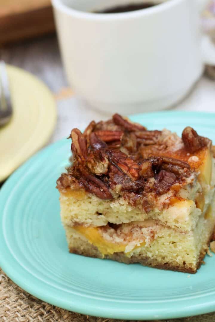 A square slice of peach coffee cake topped with pecans sits on a light blue plate, with a cup of coffee in the background.