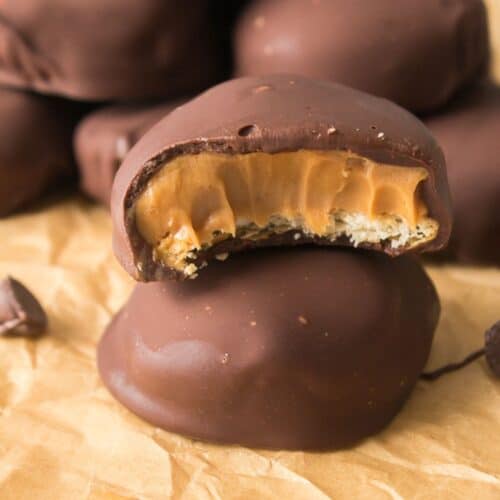 A stack of chocolate-covered Peanut Butter Patties, with one showing a bite revealing a caramel and biscuit filling, placed on brown parchment paper.