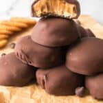 A stack of chocolate-covered cookies with a creamy peanut butter filling, one with a bite taken out, displayed on parchment paper alongside chocolate chips and crackers.