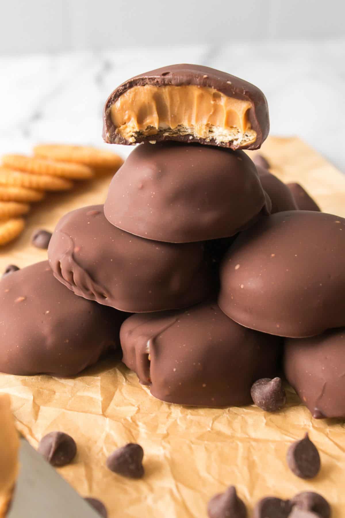 A stack of chocolate-covered cookies with a creamy peanut butter filling, one with a bite taken out, displayed on parchment paper alongside chocolate chips and crackers.