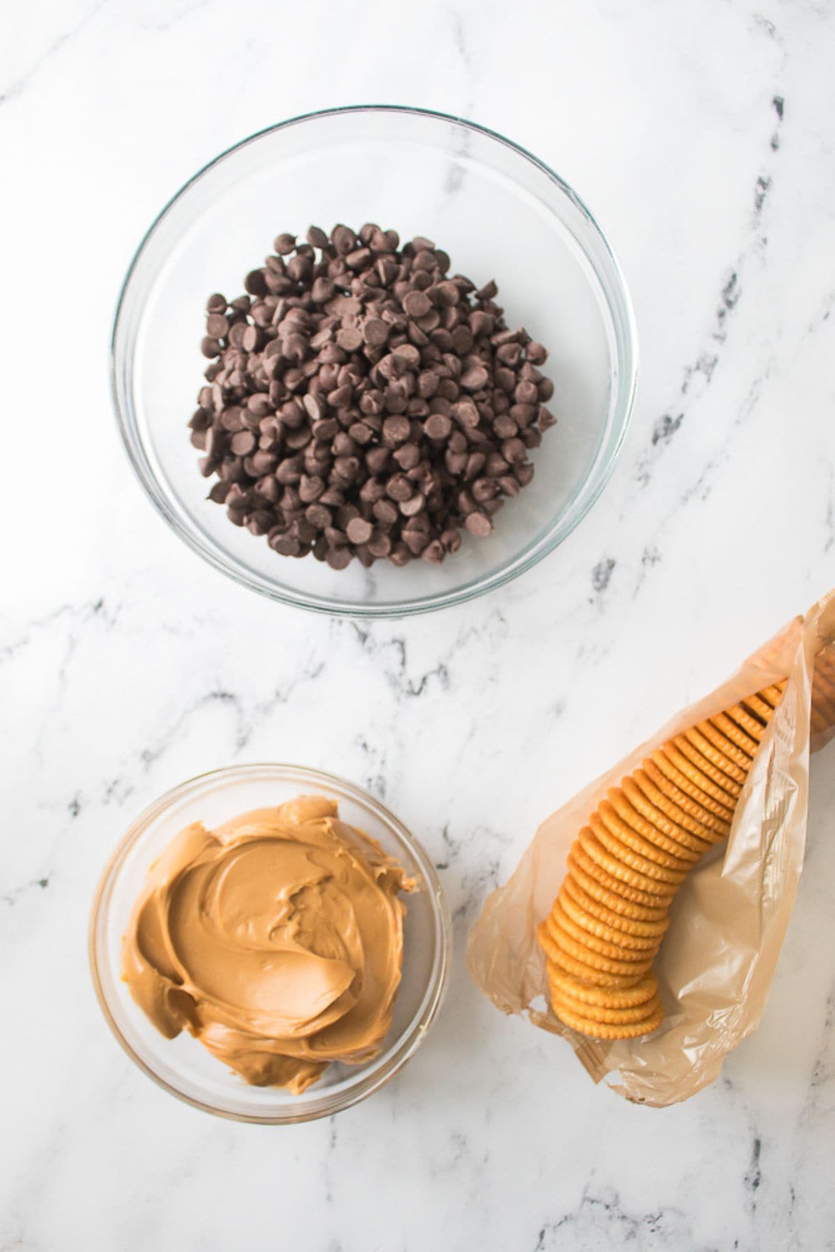 A glass bowl of chocolate chips, a glass bowl of peanut butter, and a sleeve of round crackers on a white marble surface—perfect for making homemade Peanut Butter Patties.