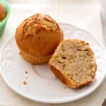 A whole Pumpkin Muffins with Spice Cake and a halved muffin are displayed on a white plate, showing the moist, spiced texture inside. Crumbs are scattered on the plate.