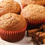 Four Pumpkin Muffins with Spice Cake flavors in red paper liners sit on a white plate, accented by cinnamon sticks and star anise.