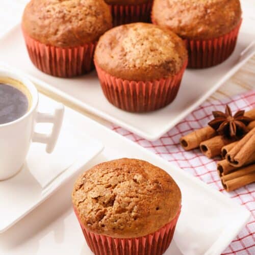 A plate of five Pumpkin Muffins with Spice Cake in red wrappers, a white cup of black coffee, and a single muffin with cinnamon sticks and star anise rest on a checkered cloth.