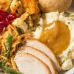 A plate with slices, mashed potatoes with gravy, green bean casserole, stuffing, cranberry sauce, and a dinner roll.