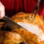 A person in a red sweater is carving a slow roasted turkey with a knife and fork.