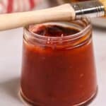 A glass jar filled with reddish-brown liquid sits on a white surface, with a basting brush resting across the top of the jar.