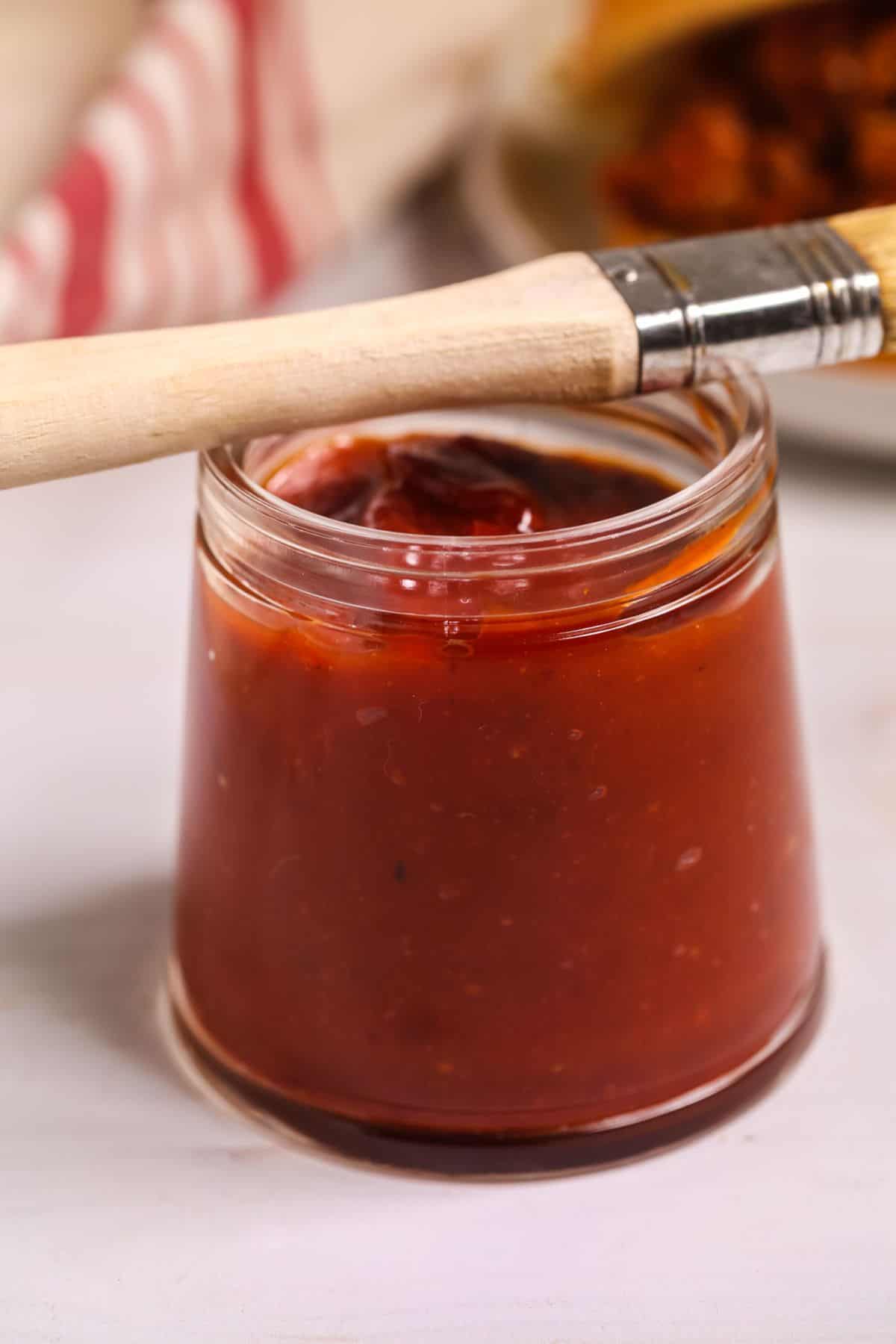 A glass jar filled with reddish-brown liquid sits on a white surface, with a basting brush resting across the top of the jar.