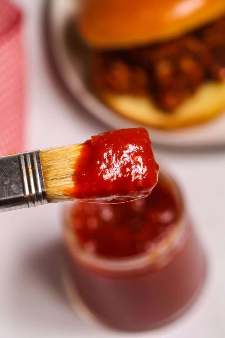 A close-up of a basting brush dipped in Chipotle BBQ Sauce, with an open jar of sauce and a sandwich bun in the background.