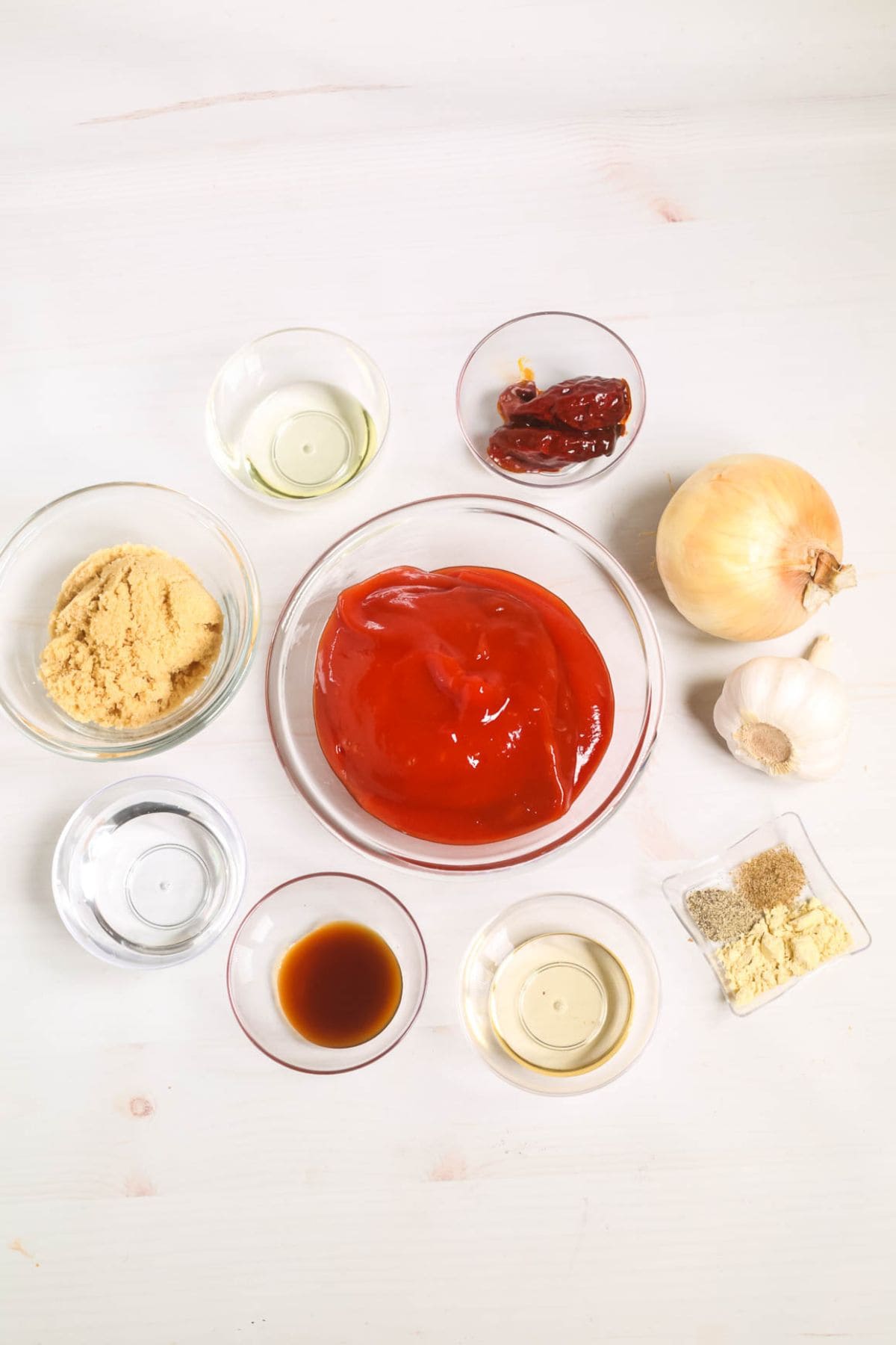 A variety of ingredients, including ketchup, brown sugar, onion, garlic, spices, vinegar, soy sauce, chili peppers, and oil, arranged in bowls on a white surface.