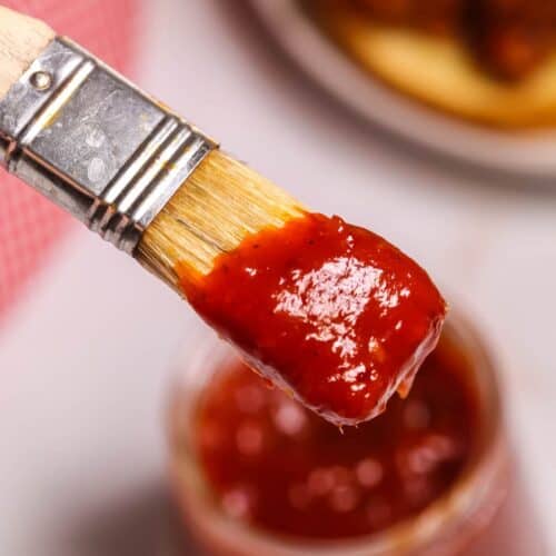 A close-up of a basting brush coated in smoky Chipotle BBQ Sauce, held above an open jar of sauce, with blurred food in the background.