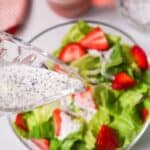 A glass pitcher pours poppy seed dressing onto a Strawberry Poppyseed Salad with fresh lettuce and sliced strawberries in a clear bowl.