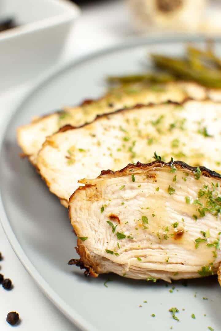 Close-up of sliced air fryer turkey breast garnished with chopped herbs, served on a gray plate with green beans in the background.