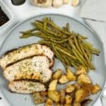 A plate with sliced Air Fryer Turkey Breast, roasted potatoes, and cooked green beans, set on a white surface with garlic, peppercorns, and a gray napkin nearby.