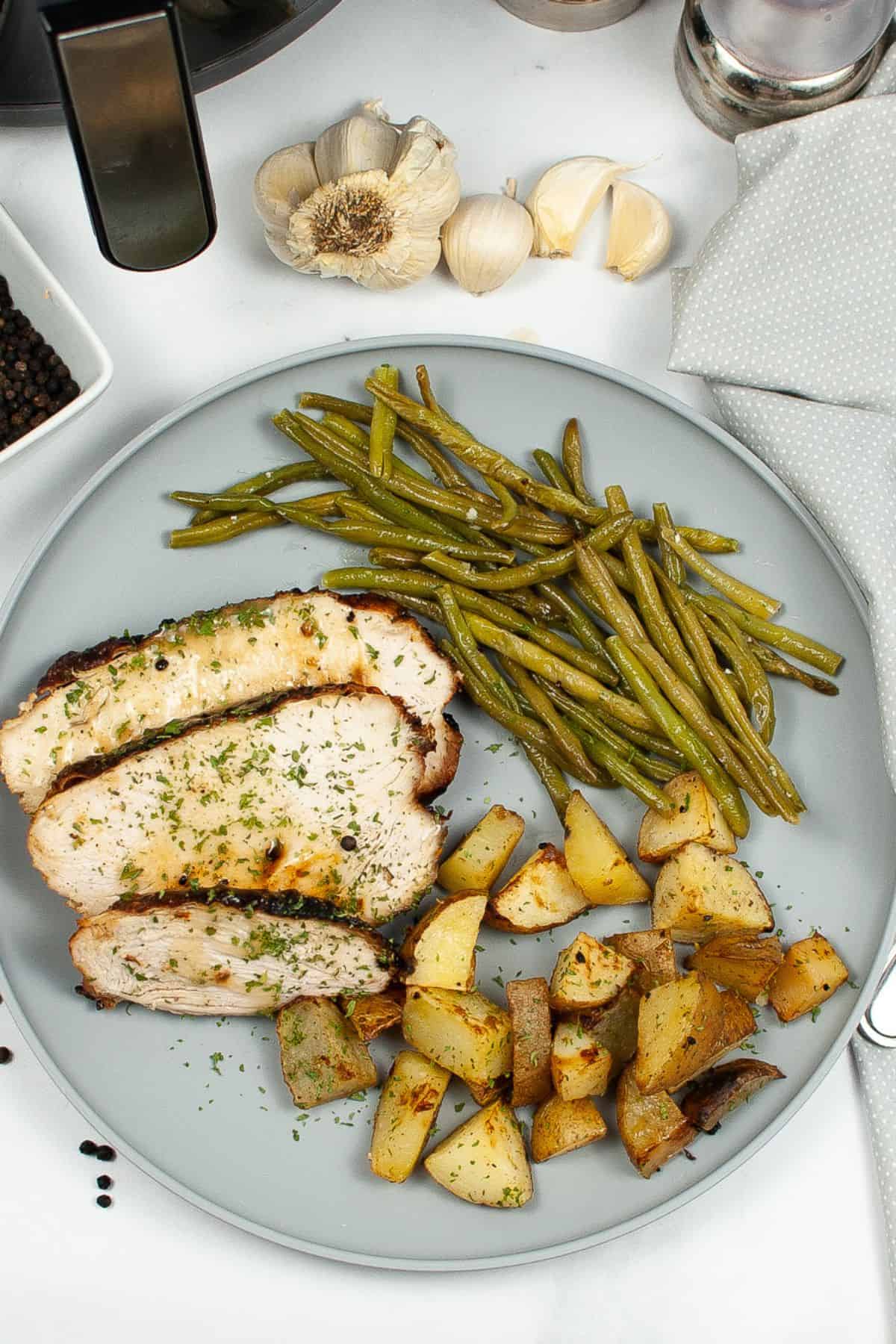A plate with sliced Air Fryer Turkey Breast, roasted potatoes, and cooked green beans, set on a white surface with garlic, peppercorns, and a gray napkin nearby.