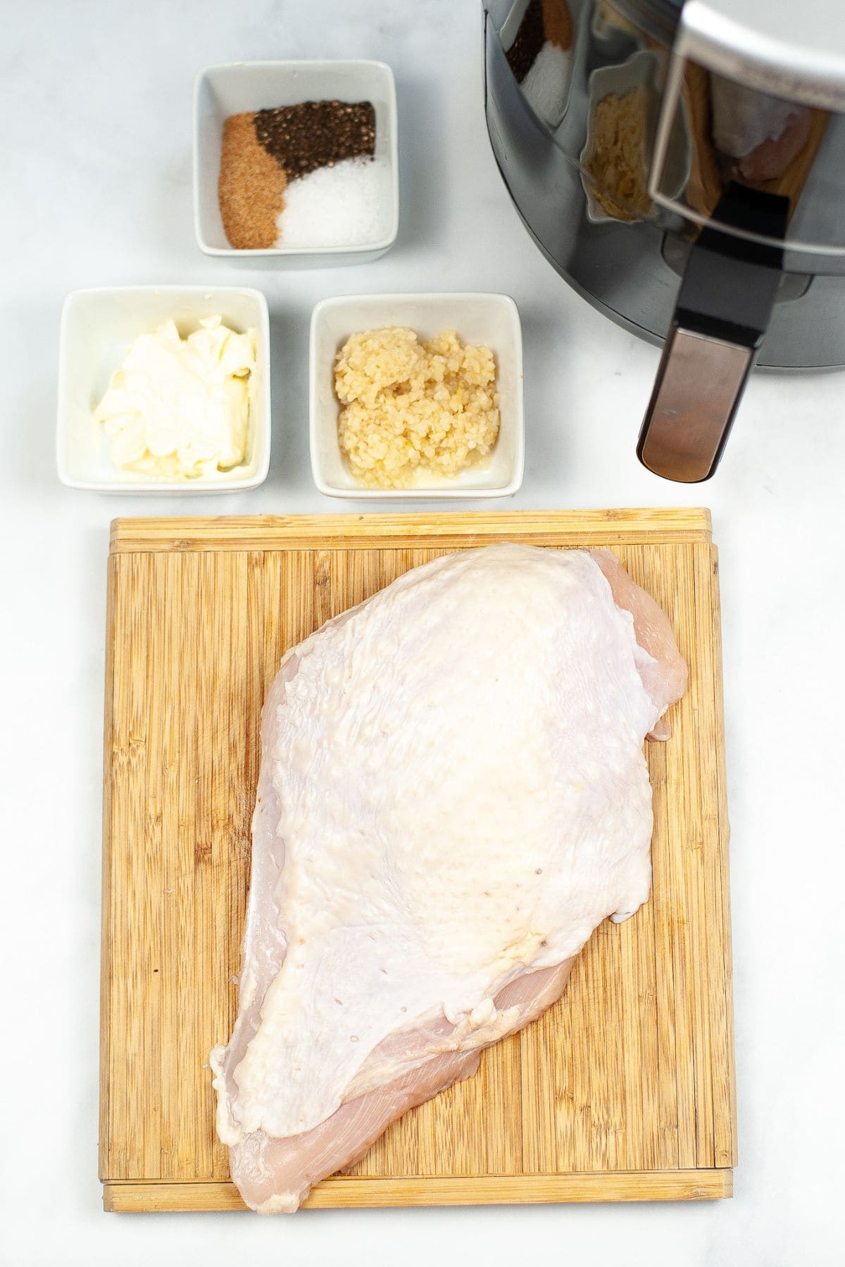 Raw turkey breast on a wooden cutting board with bowls of spices, garlic, and butter nearby, perfect for preparing Air Fryer Turkey Breast next to an air fryer on a white surface.