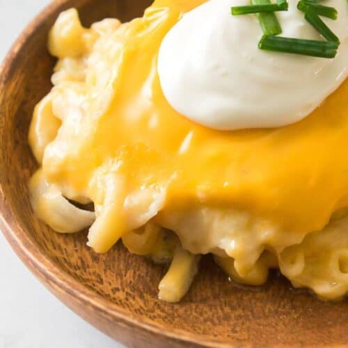 A bowl of cheesy pasta reminiscent of a Baked Pierogi Casserole, topped with a luscious slice of melted cheese, tangy sour cream, and a sprinkle of chopped chives on a rustic wooden surface.