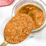 A close-up of a spoonful of burger seasoning held above an open jar filled with the same reddish-brown spice blend. A red and white checkered fabric is visible in the background.