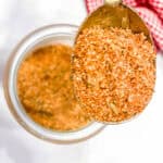 A spoonful of orange-red burger seasoning mix held above an open glass jar filled with the same spice, with a red and white checkered fabric in the background.