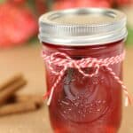 A glass jar filled with red Cranberry Moonshine, sealed with a metal lid and decorated with a red and white string, sits on a surface with cinnamon sticks and red decorations in the background.