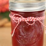 A glass jar filled with red cranberry moonshine jam, sealed with a metal lid and decorated with a red and white twine bow.