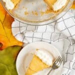 A slice with whipped cream on a plate and fork, next to the remaining pie in a dish; autumn leaf and kitchen towel beside them.