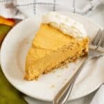 A slice of Cream Cheese Pumpkin Pie cheesecake with a graham cracker crust and whipped cream on top, served on a white plate with a fork.