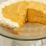 A creamy dessert sits on a glass cake stand, with one slice missing and whipped cream piped around the edge.