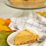 A slice of Cream Cheese Pumpkin Pie with whipped cream on a white plate and fork, with the remaining pie on a glass stand in the background.