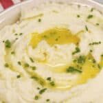 A bowl of whipped potatoes topped with melted butter and chopped chives, with a spoon and striped napkin in the background.