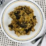 A plate of Crock Pot Stuffing with pieces of mushrooms sits on a white plate with a dotted border, surrounded by patterned napkins and two forks.