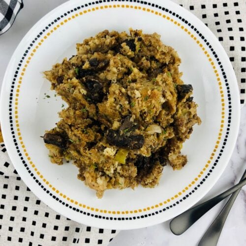A plate of Crock Pot Stuffing with pieces of mushrooms sits on a white plate with a dotted border, surrounded by patterned napkins and two forks.