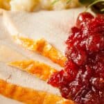 Close-up of sliced Crock Pot Turkey Breast with golden skin, topped with cranberry sauce, mashed potatoes, gravy, and a sprig of rosemary.